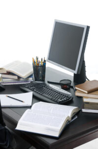 Schreibtisch mit offenen Büchern, Stiften, Schreibblock, Computermonitor und Tastatur.