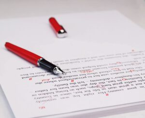 A ballpoint pen lies on a printout of text with various corrections made in red.
